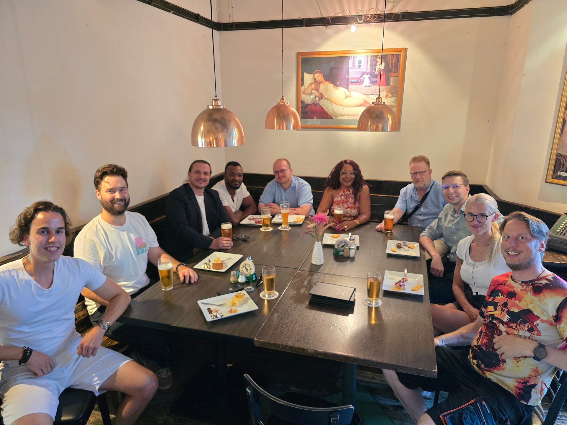 Gruppenfoto vom KI-Stammtisch im Juli 2026 — zehn Teilnehmende an einem langen Tisch im UNIKUM Augsburg