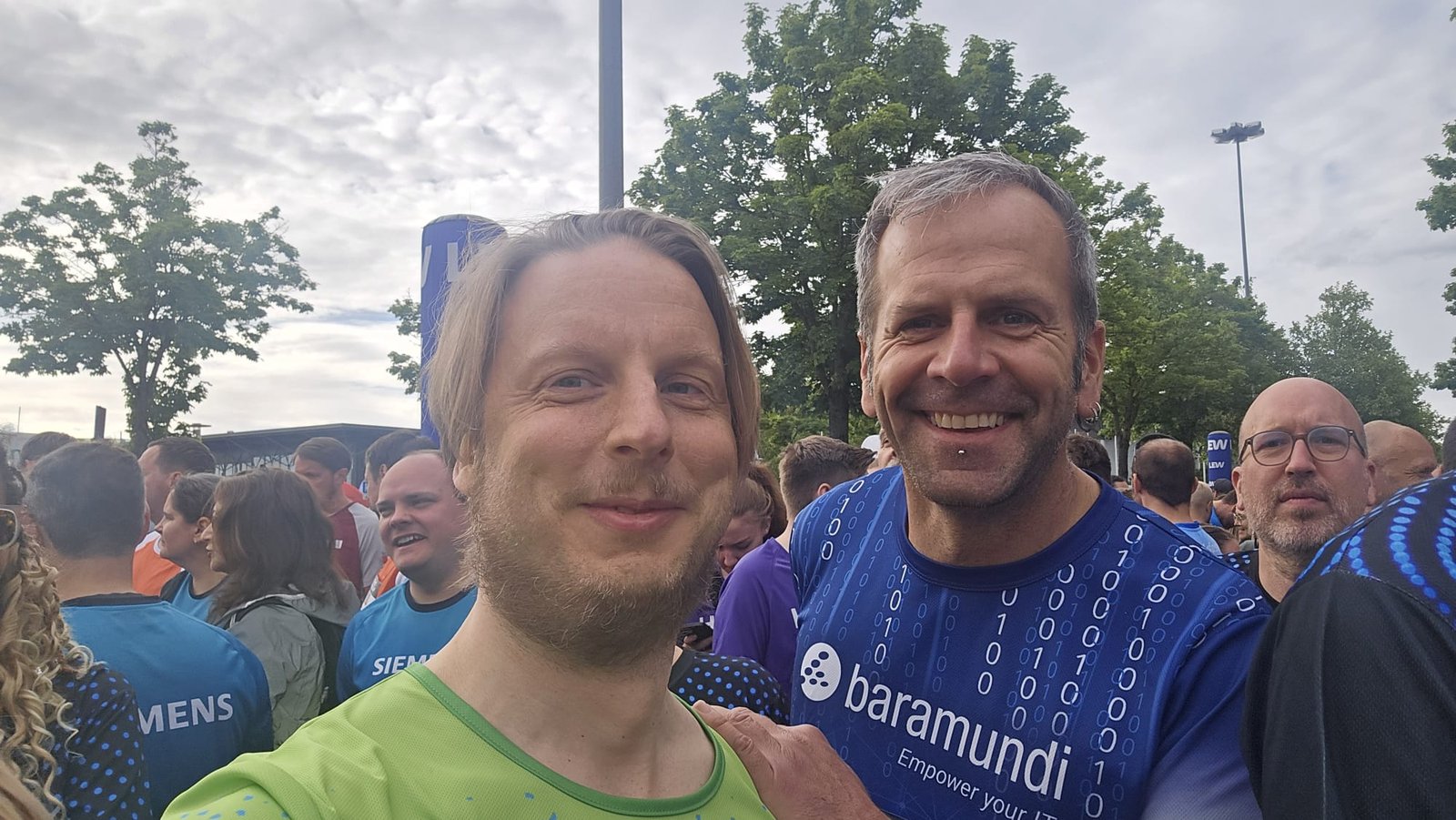 Florian Ludwig (links) im Startbereich des M-net Firmenlaufs Augsburg 2026, daneben ein Mitläufer im Firmen-Laufshirt — im Hintergrund hunderte Läuferinnen und Läufer in Team-Trikots.