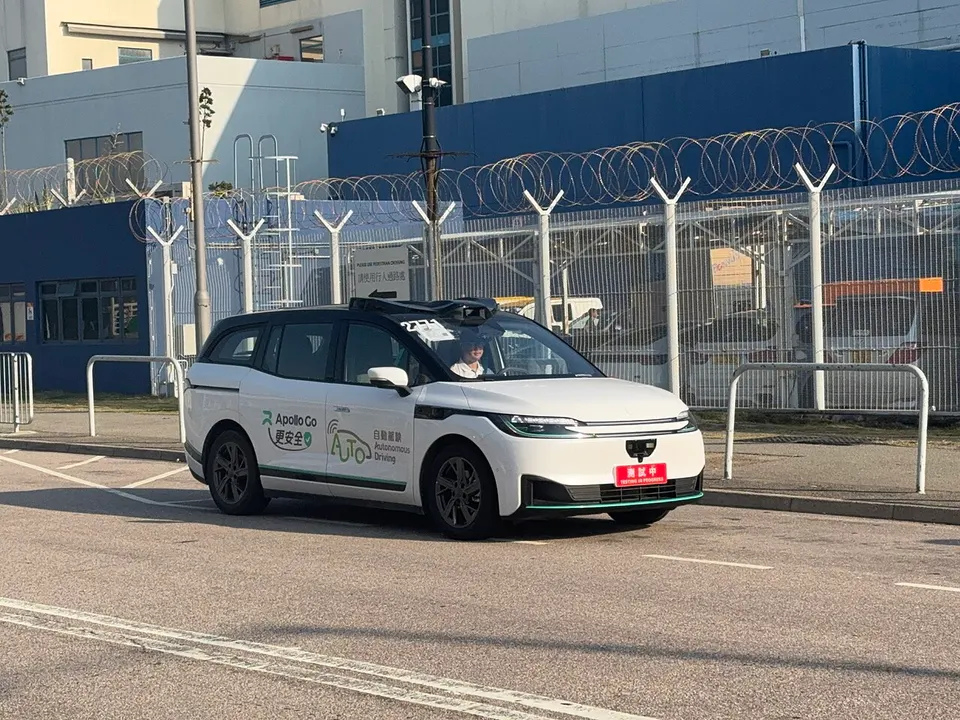 Weißes Robotaxi mit Dachsensoren auf einer leeren Betriebsstraße vor einem Zaun mit Stacheldraht