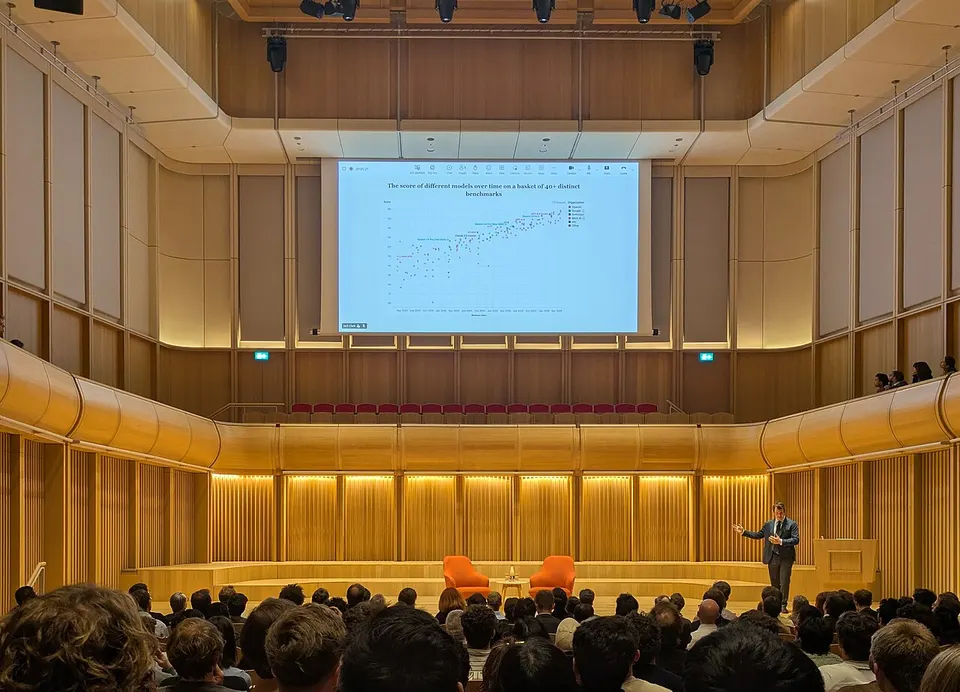 Vortragssaal mit holzverkleideten Wänden, auf der Leinwand ein Punktediagramm mit steigenden Benchmark-Werten, rechts unten ein Redner