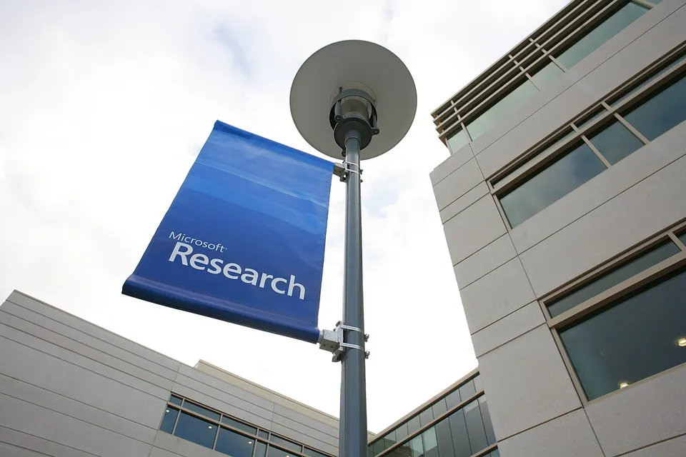 Laternenmast mit blauem Banner „Microsoft Research“ vor einem modernen Bürogebäude mit Glasfassade