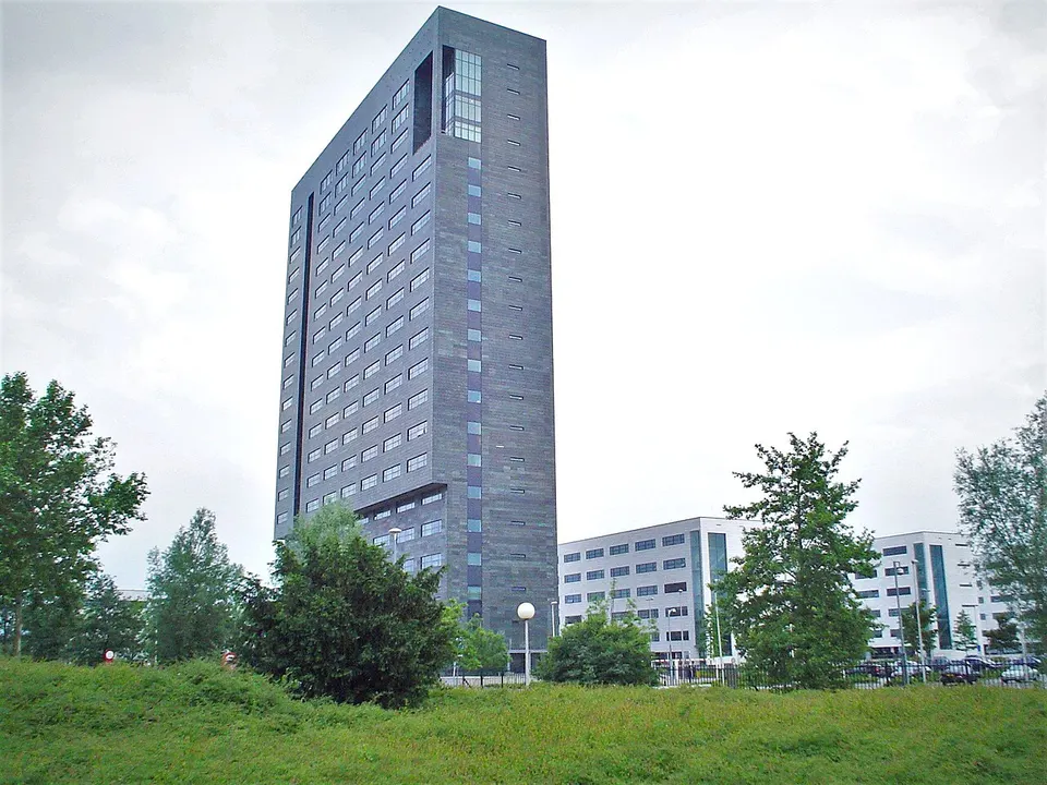 Schmales, dunkelgraues Bürohochhaus hinter Bäumen und einer Wiese, rechts flachere weiße Bürogebäude