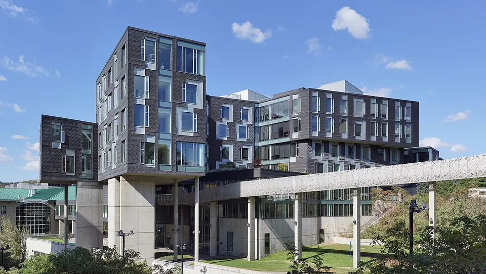 Modernes Gebäude mit dunkler Schuppenfassade und versetzten Fenstern, davor eine Fußgängerbrücke und Rasen